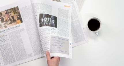 Iemand die een papieren krant vasthoudt en een kopje koffie naast zich heeft staan
