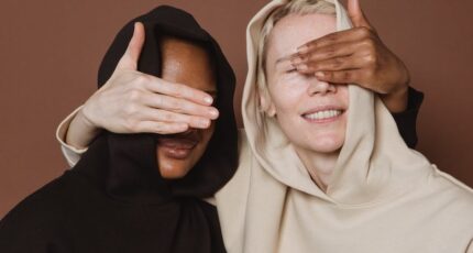 Content diverse vrouwelijke modellen in casual hoodies die elkaars ogen bedekken met hun handen terwijl ze in de studio op een bruine achtergrond staan