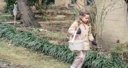 Meisje in warme kleding draagt stoffen mand en zoekt naar verstopte paaseieren terwijl ze loopt in weelderige lentetuin