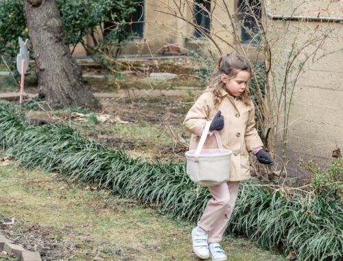 Meisje in warme kleding draagt stoffen mand en zoekt naar verstopte paaseieren terwijl ze loopt in weelderige lentetuin