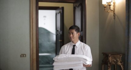 Jonge mannelijke huishoudelijke werknemer draagt een stapel witte beddengoed terwijl hij een hotelkamer binnenkomt.