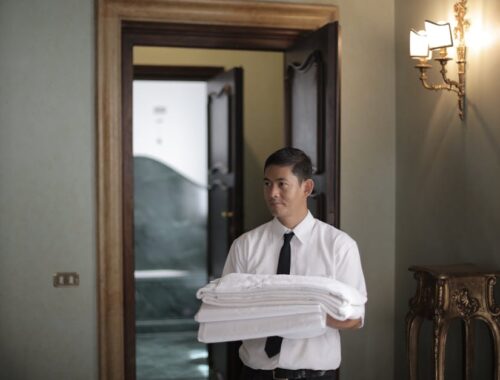 Jonge mannelijke huishoudelijke werknemer draagt een stapel witte beddengoed terwijl hij een hotelkamer binnenkomt.