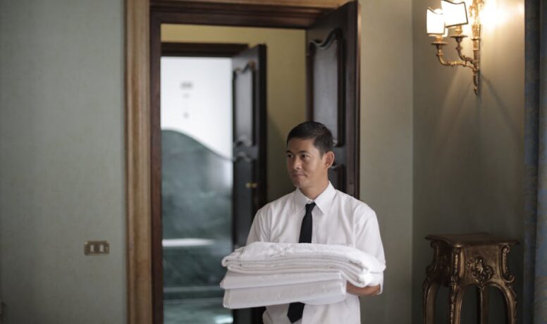 Jonge mannelijke huishoudelijke werknemer draagt een stapel witte beddengoed terwijl hij een hotelkamer binnenkomt.