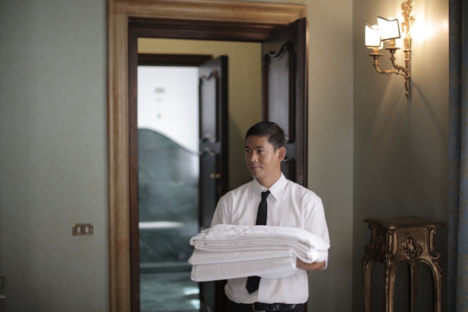 Jonge mannelijke huishoudelijke werknemer draagt een stapel witte beddengoed terwijl hij een hotelkamer binnenkomt.