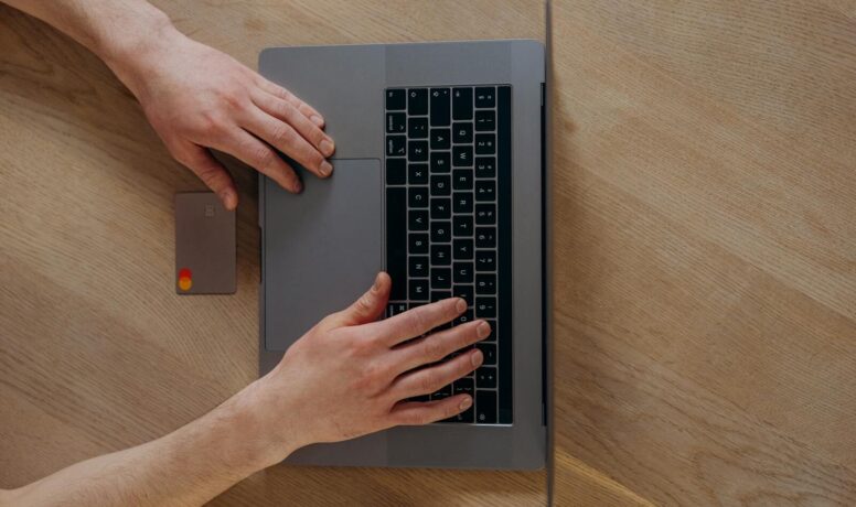 A person using a laptop and credit card for online shopping, viewed from above on a wooden table.